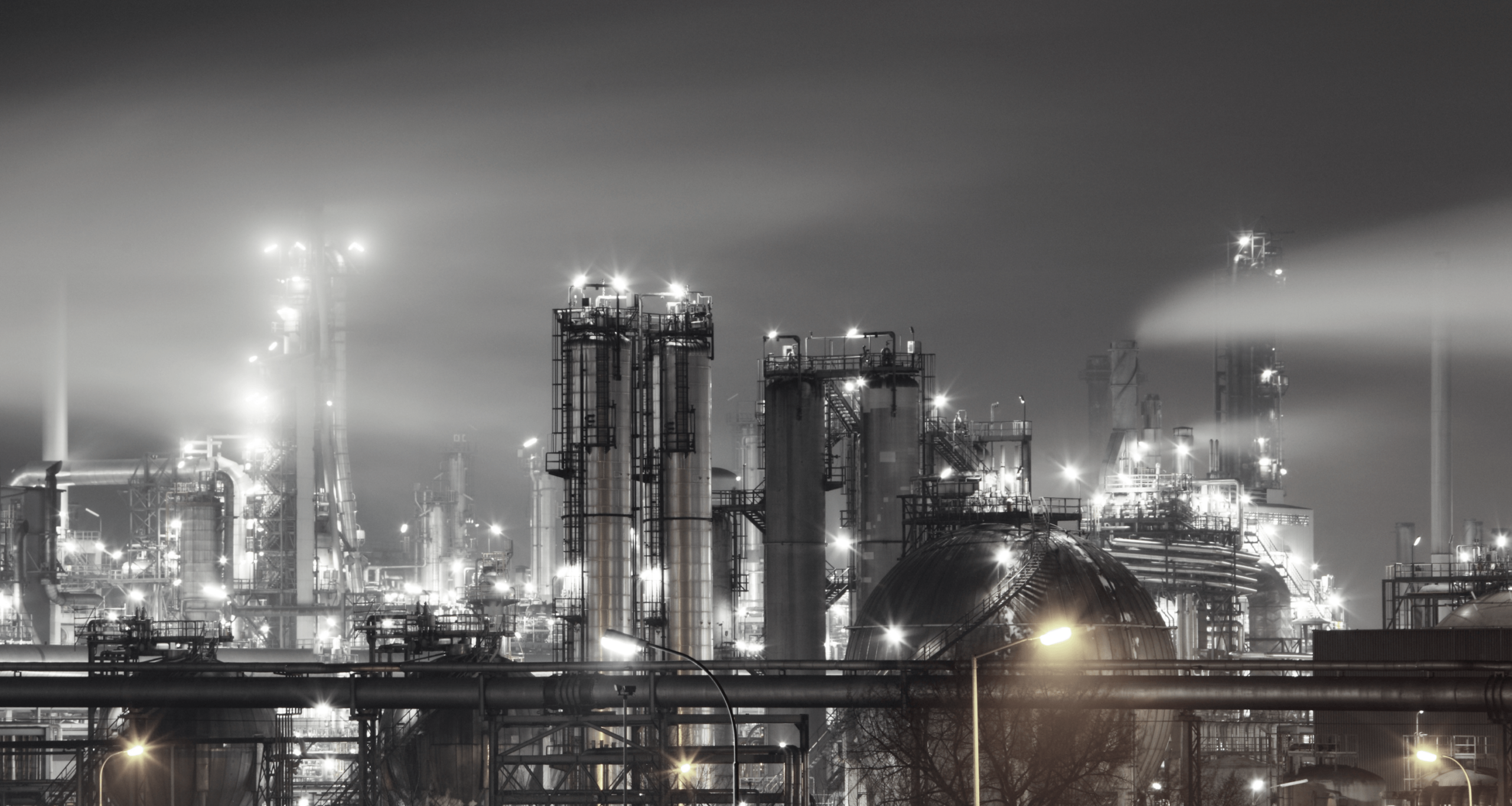 Industrieanlage bei Nacht mit beleuchteten Türmen, Schornsteinen und Rohrleitungen, aufgenommen in Schwarz-Weiß.